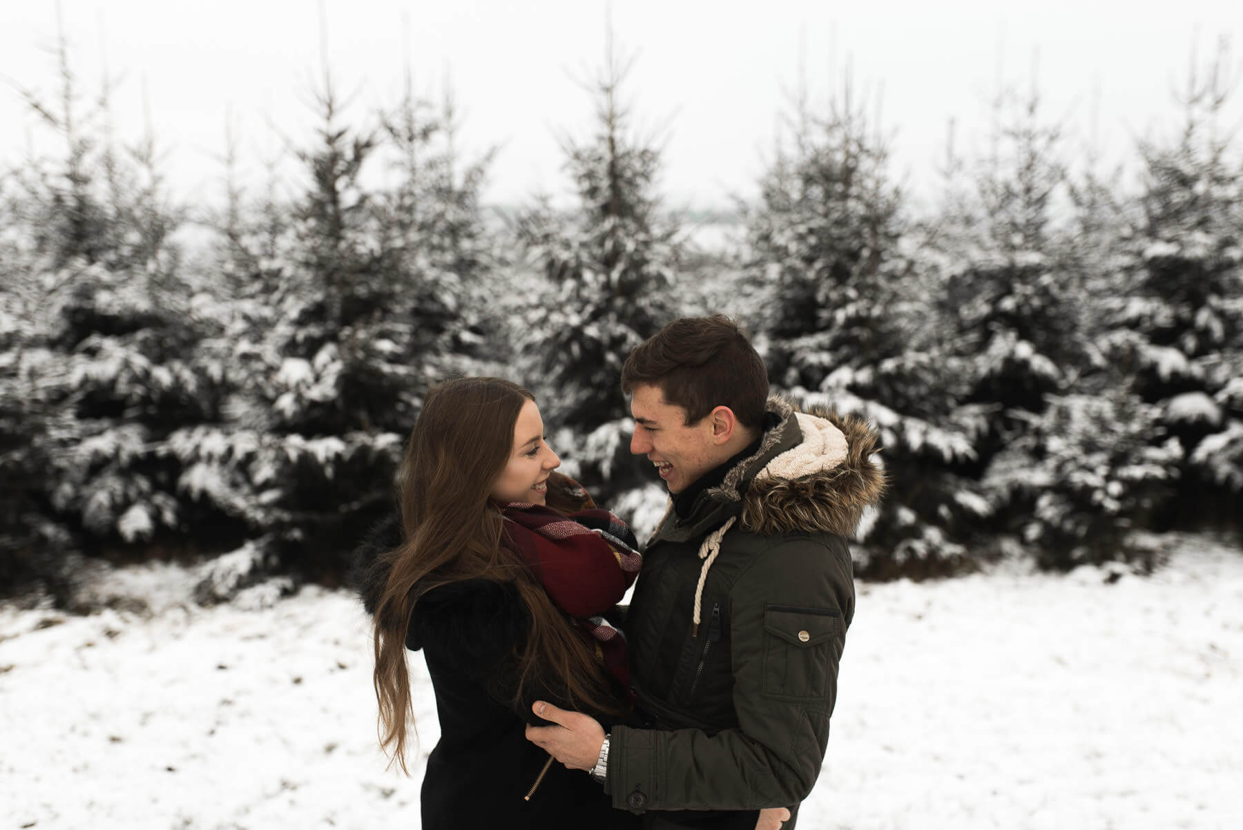 Verliebtes Paar lachend im Schnee vor Nadelwald mit Umarmung