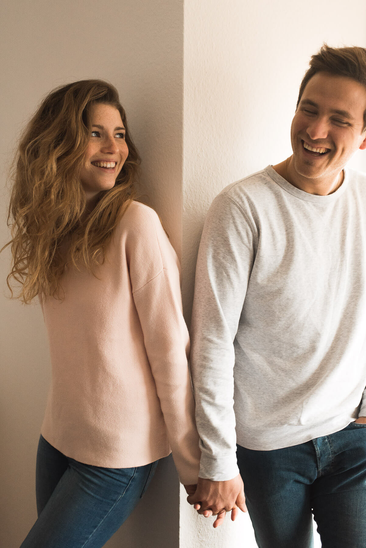 Paar steht lachend in den eigenen vier Wänden vor weißer Wand mit Pullover und schaut sich an