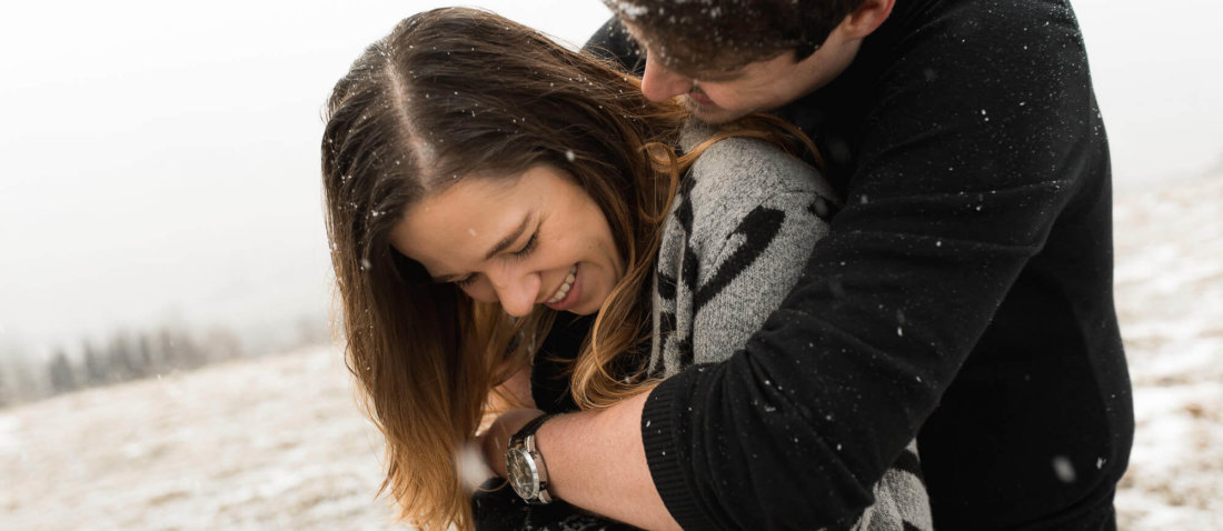 Mann umarmt seine Frau und sie lacht dabei während sie im Schnee stehen