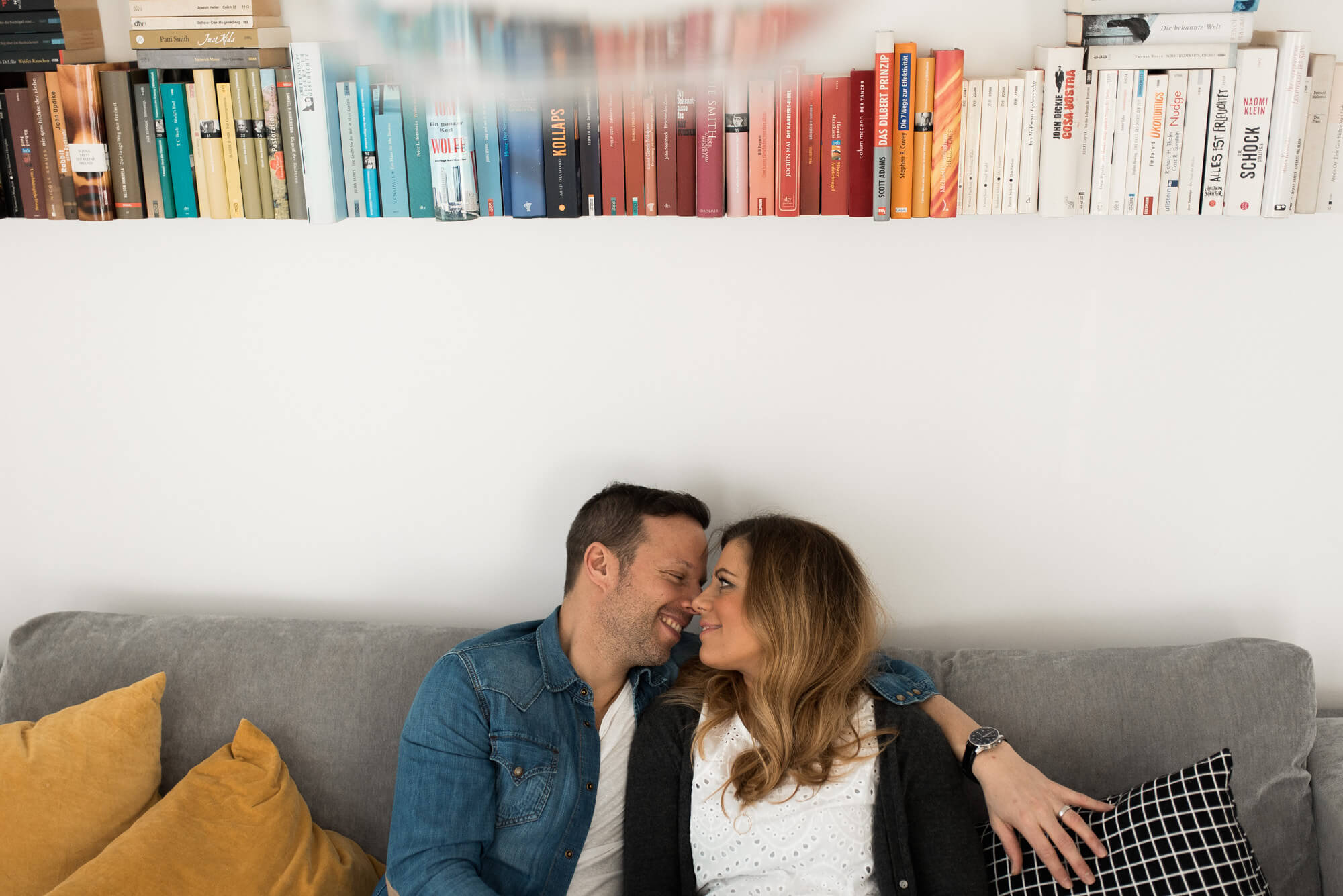 Paar hockt auf Couch unter Bücherregal und lächelt