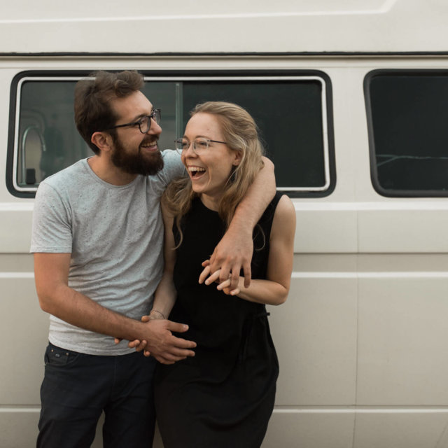 Lachendes Paar steht vor ihrem Volkswagen Bully Hand in hand