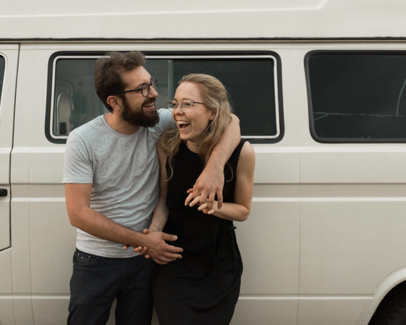 Lachendes Paar steht vor ihrem Volkswagen Bully Hand in hand