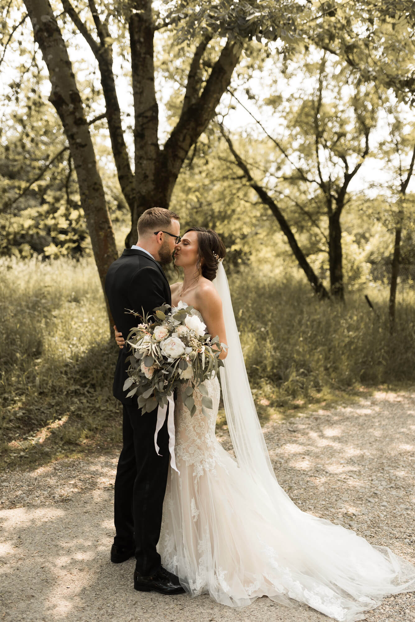 Brautpaar steht im Wald bei Sonne und küsst sich mit einem Brautstrauss von My Vivent mit Eukalyptusstrauss