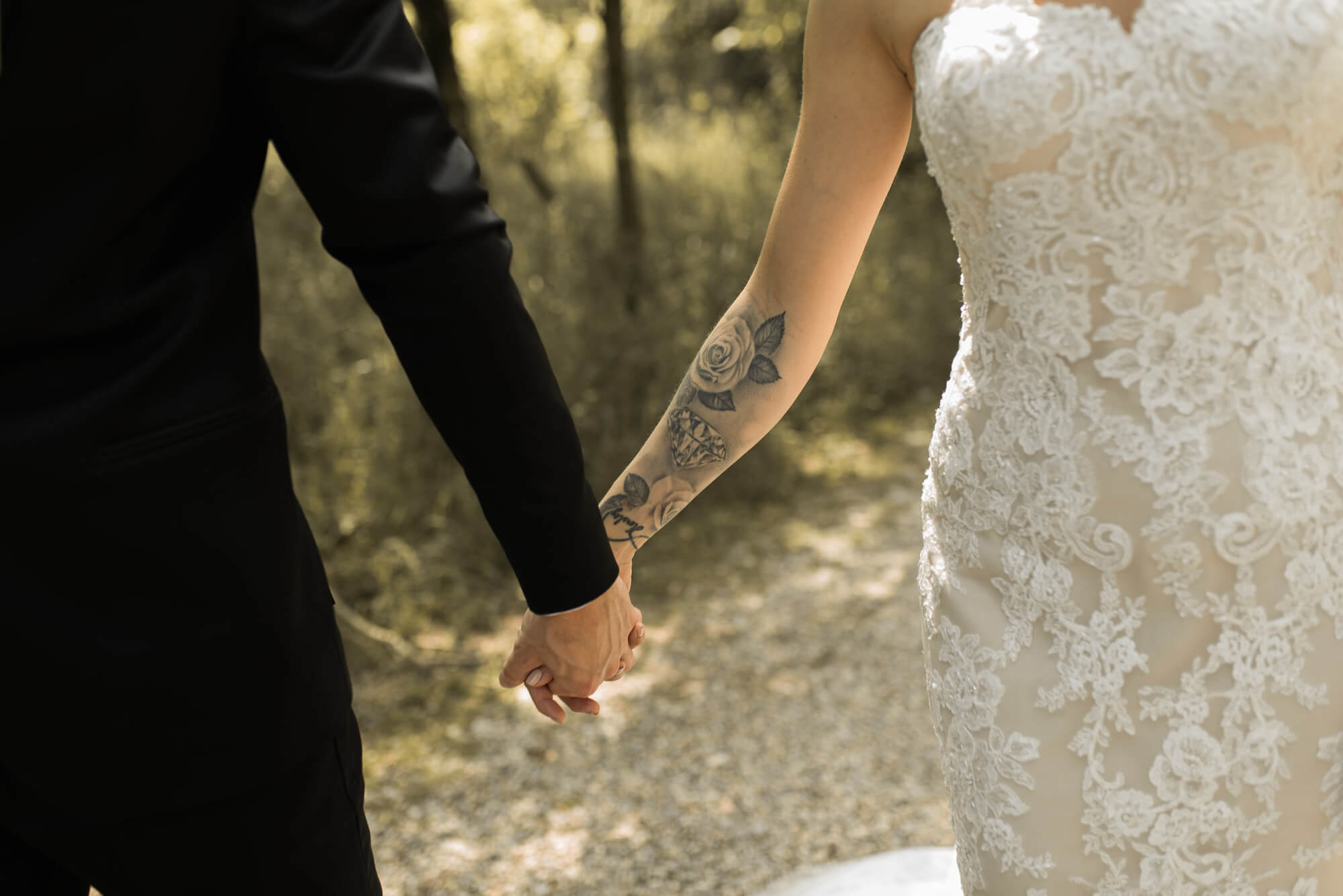 Nahaufnahme von Brautpaar die Hand in Hand durch Park laufen dabei ist ein Tattoo am Unterarm der Braut zu sehen