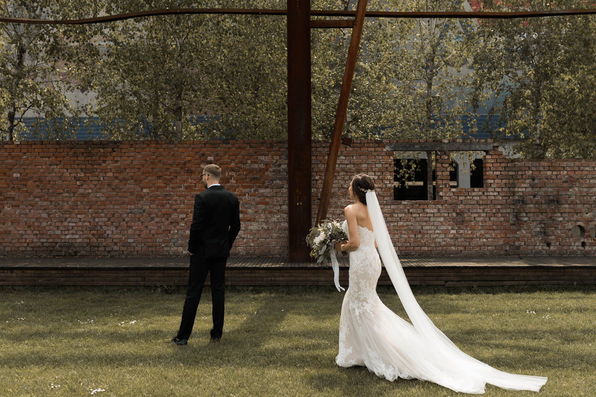 Braut mit Hochzeitskleid und Schleier läuft bei First Look auf den Bräutigam zu der mit dem Rücken zu ihr auf einer Wiese vor Ziegelsteinmauer