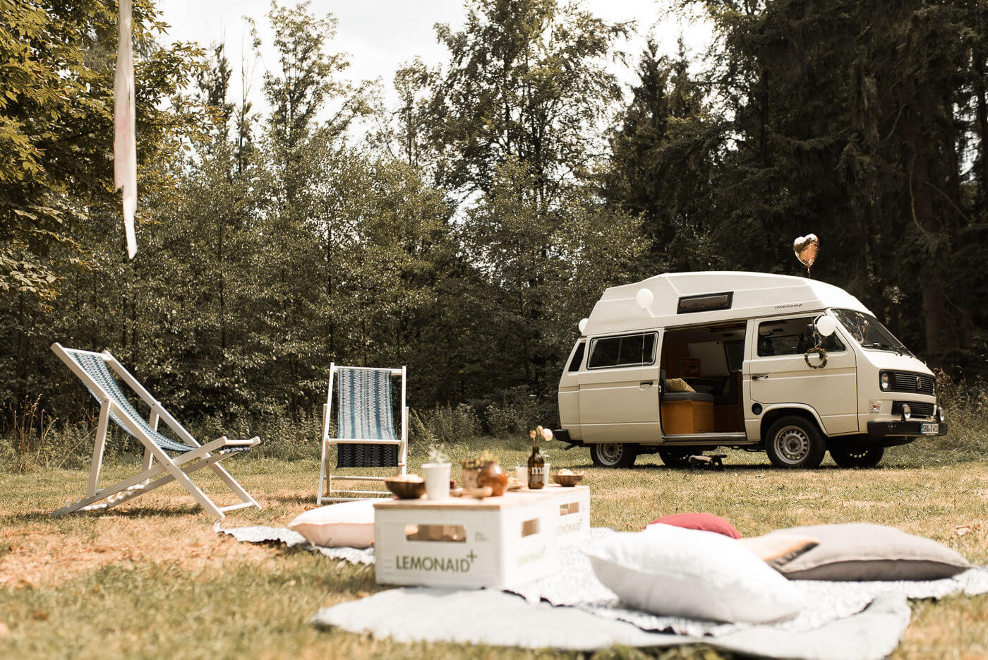 Sektempfang Deko mit Teppich und Sitztischen auf Waldwiese mit weißen Volkswagen Bully als Hochzeitsauto