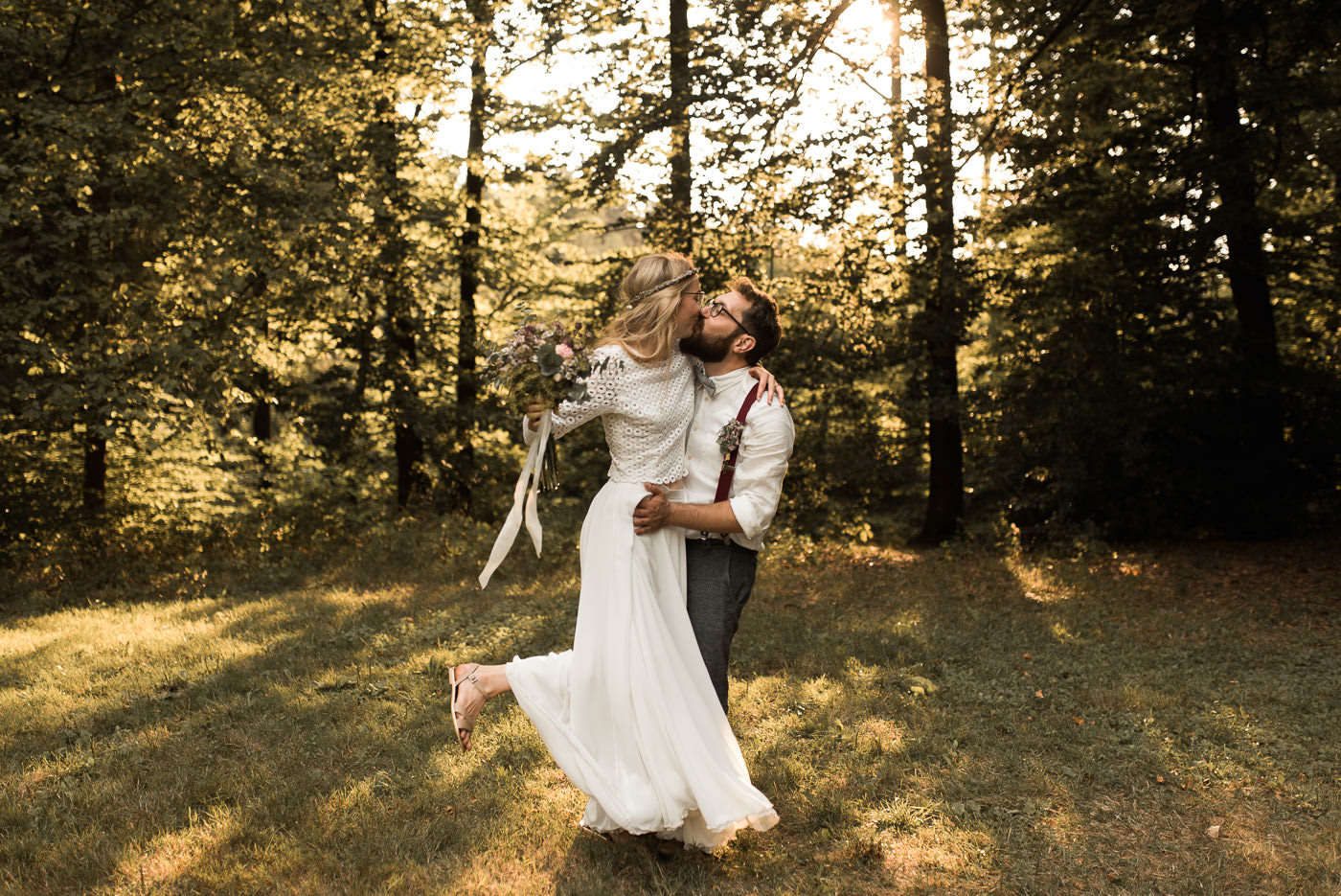 Braut springt Mann in die Arme und küssen sich am Hochzeitstag im Wald bei Sonne auf grüner Wiese