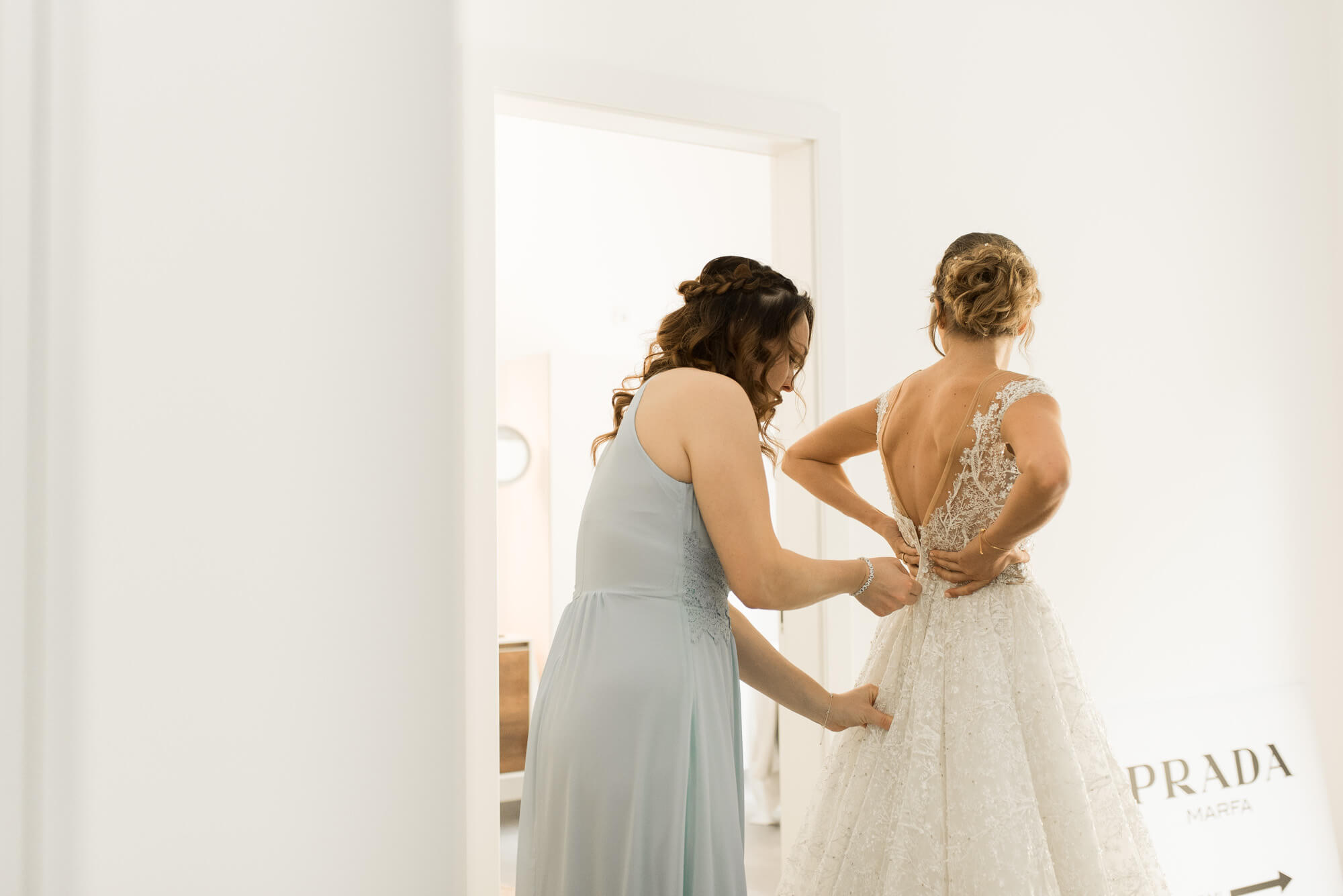 Trauzeugin schließt der Braut das Hochzeitskleid von Lorenzo Rossi und im Hintergrund ist ein Schild von Prada