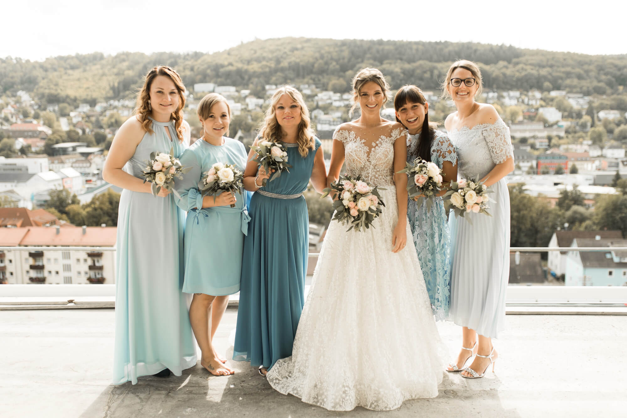 Braut steht mit Brautjungfern die ihr blaues Kleid tragen mit Stadt Panorama im Hintergrund mit Brautstrauß Blumen von Grüner Garten Mosbach und einem Hochzeitskleid von Lorenzo Rossi